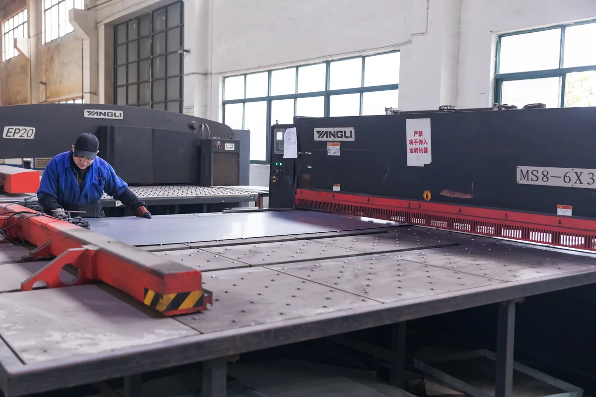 Keshun worker feeding steel sheet into a hydraulic shearing machine beside the CNC turret punch